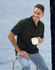 A man sits on a railing, wearing a black sustainable jacquard knit polo shirt and holding a white mug.