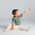 A baby wearing a green organic cotton T-shirt and cream-coloured pants, reaching out with one hand.