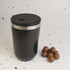 A reusable black coffee cup with a stainless steel band and a black lid, alongside a handful of chocolate balls.