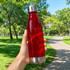 A red drink bottle with a stainless steel base and a shiny silver lid, held against a green park backdrop.