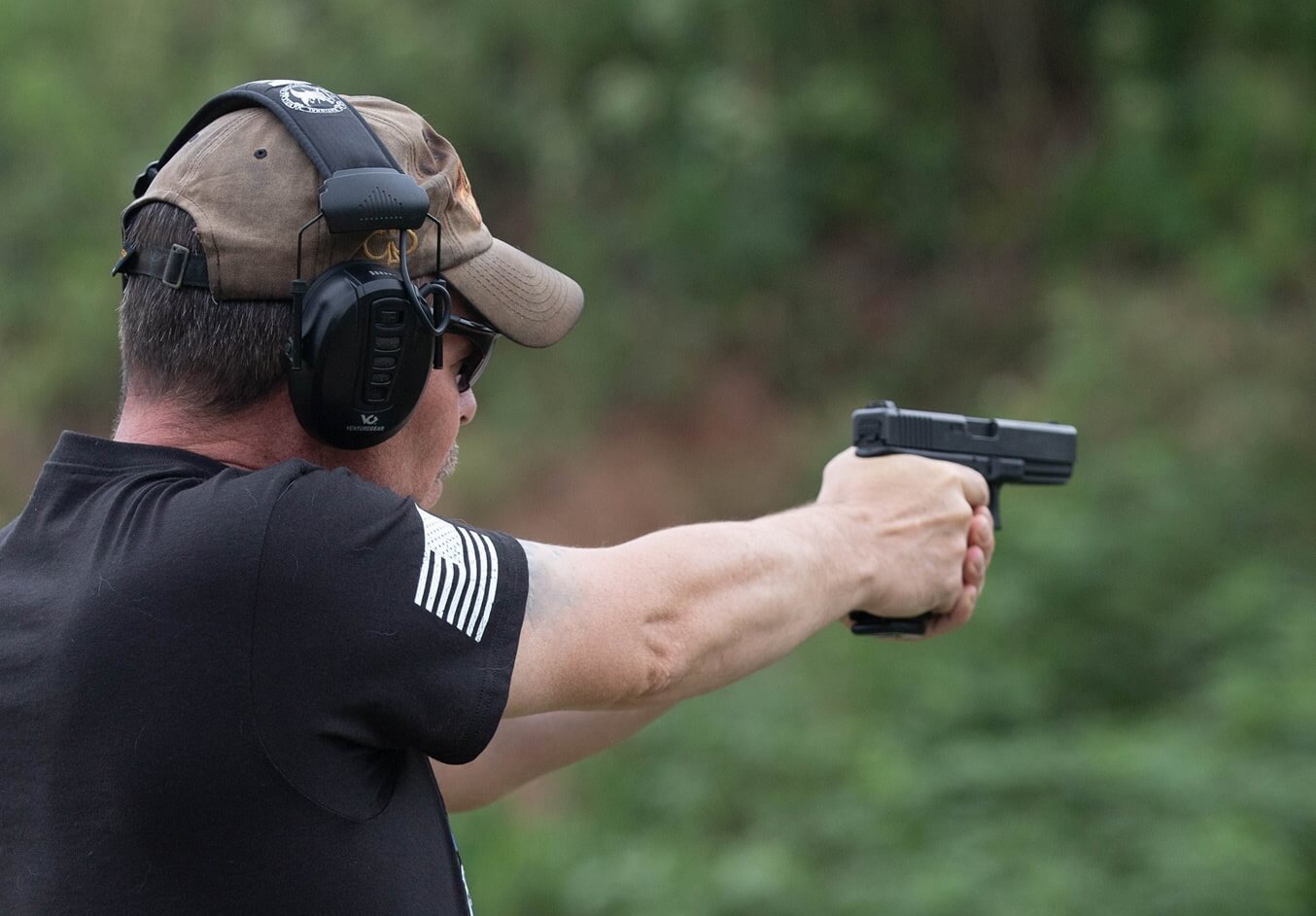 Man wearing ear protection and a cap aiming a handgun at an outdoor shooting range.