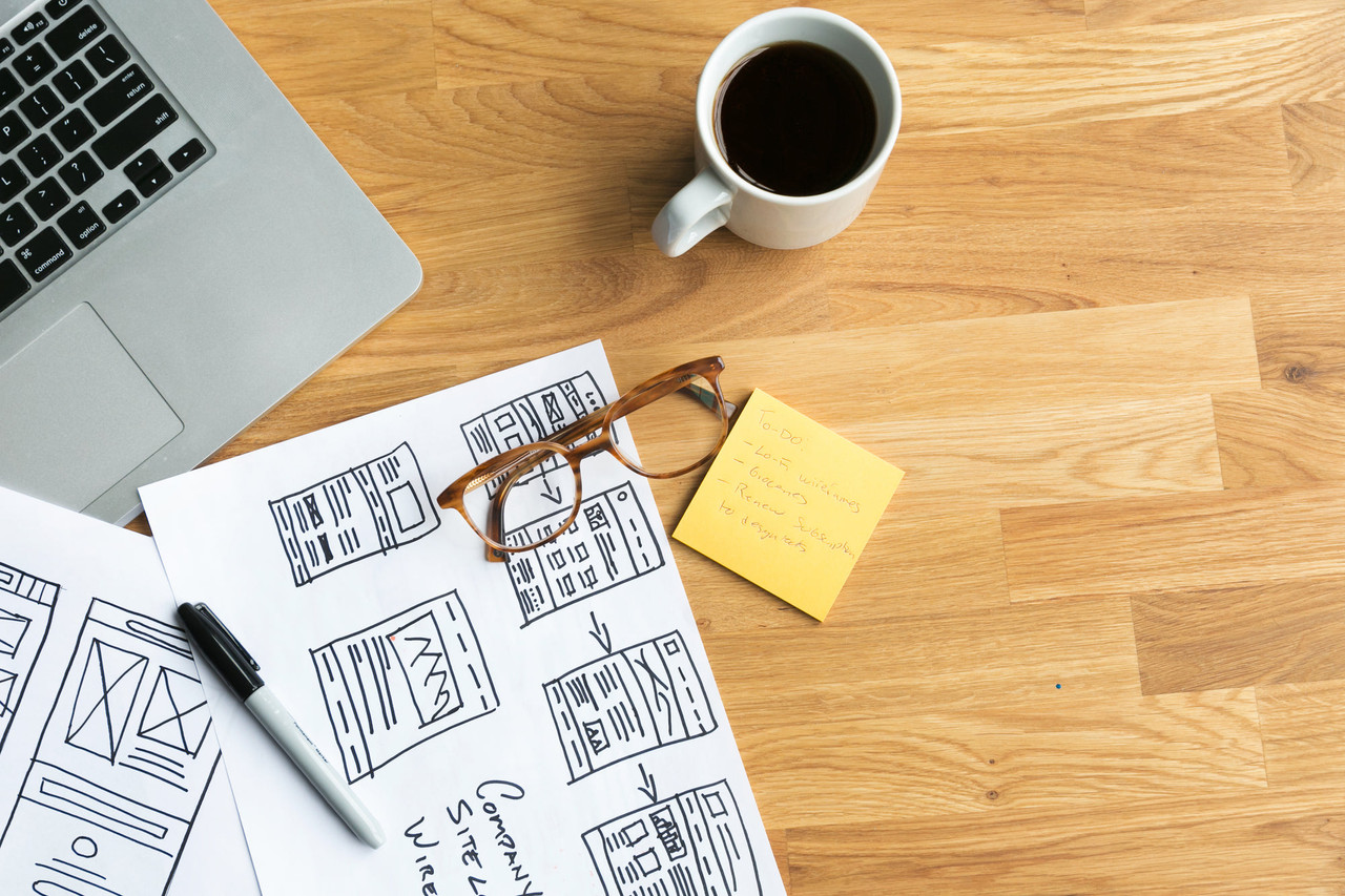 A laptop, wireframe sketches and a coffee on a desk