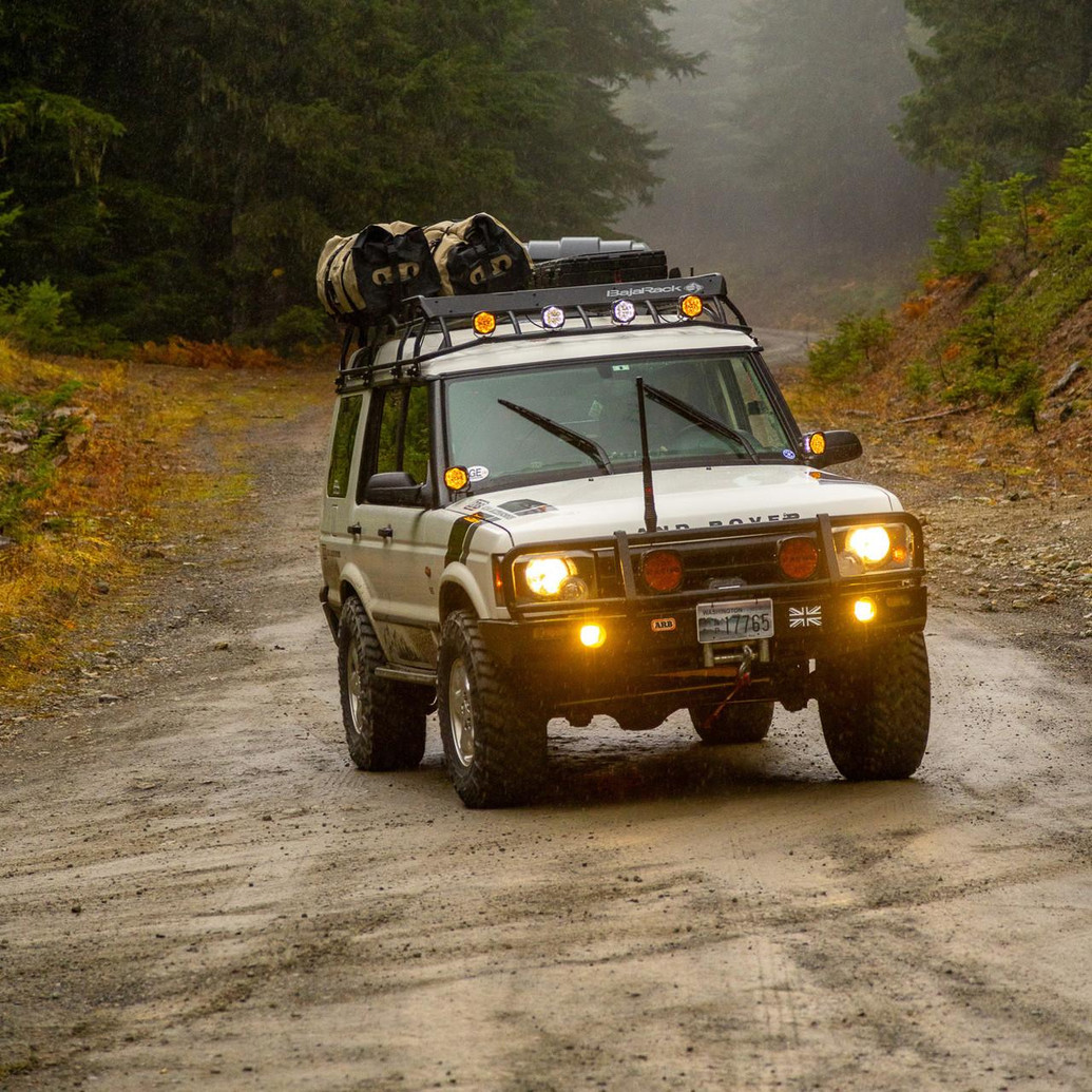 ARB Deluxe Winch Bumper 3432060B, off-road vehicle with winch bumper and roof rack, driving on a rugged dirt trail in a forested area.