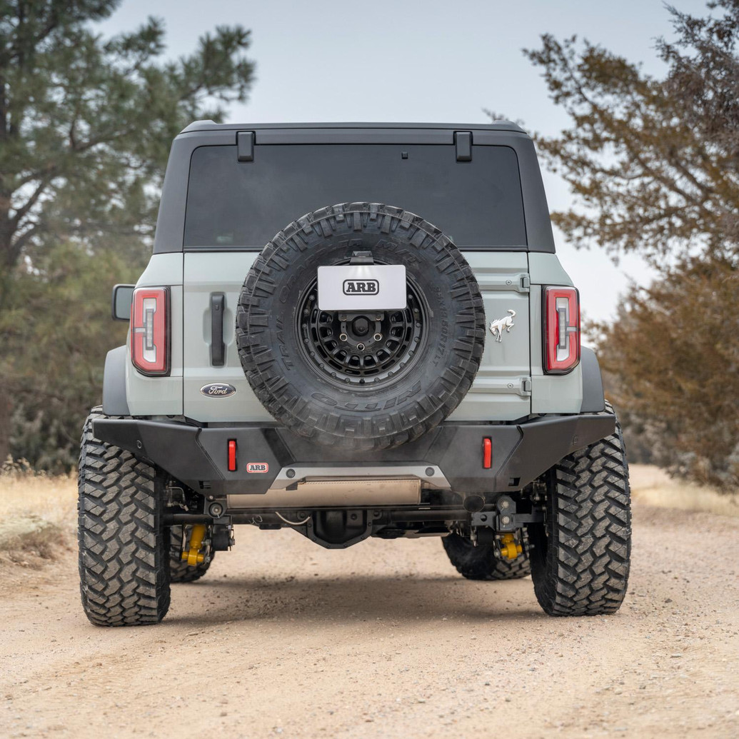 ARB Rear Zenith Bumper 5680020B, black off-road rear bumper installed on the back of a Ford Bronco with a rear-mounted spare tire, shown from behind on a dirt trail.