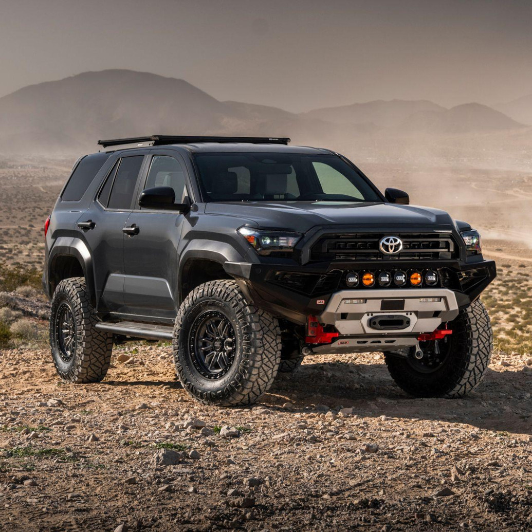 Old Man Emu MT64 Kit FR 3 in/0 lb, RR 2 in/0 lb 2134K, a rugged Toyota off-road vehicle with large tires, black grille, and additional front bumper lights, driving across a rocky desert landscape.