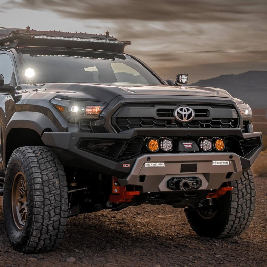 ARB Zenith Winch Bumper for Toyota Tacoma (2024+), black off-road style front bumper with integrated winch mount, four round auxiliary lights, and rugged design on a Toyota Tacoma vehicle in a desert setting.