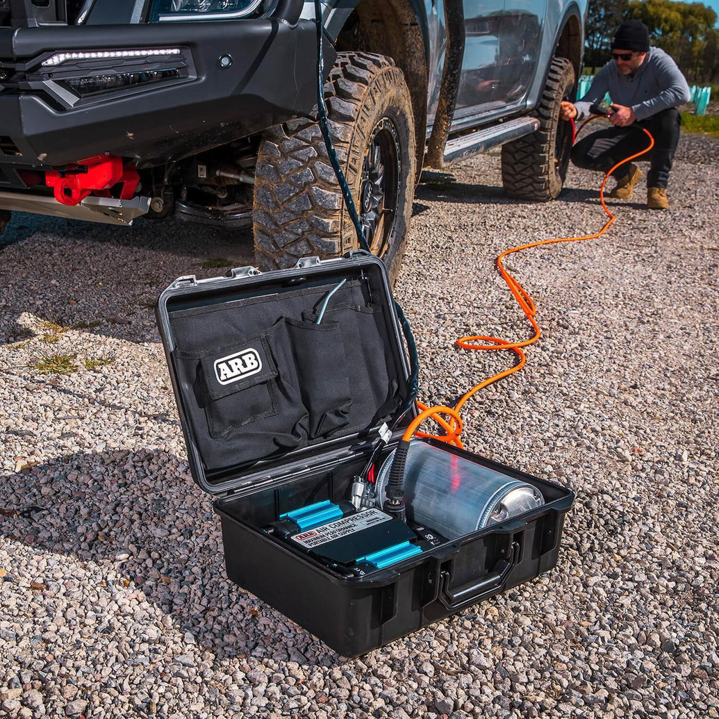 ARB Twin Motor Portable 12V Air Compressor CKMTP24, portable air compressor kit connected to an off-road vehicle for tire inflation, with a man operating the device outdoors on a gravel surface.