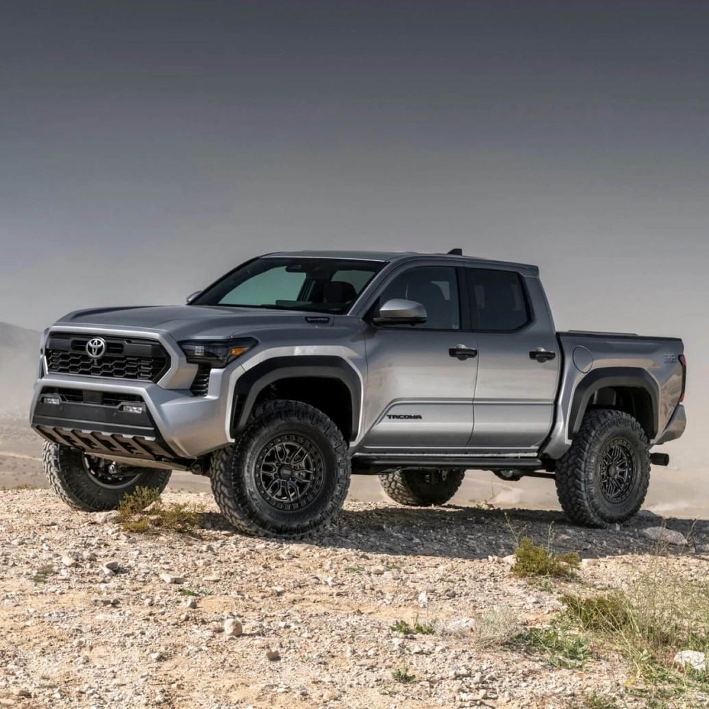 Old Man Emu MT64 Suspension Kit w/FB UCA for Tacoma 2024+, front 3 inches/0 pounds, rear 2 inches/0 pounds, installed on a silver 2024+ Toyota Tacoma pickup truck parked on rocky terrain in a desert landscape.