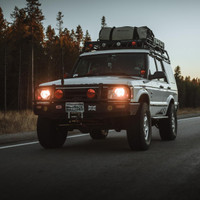 ARB Deluxe Winch Bumper 3432060B, off-road vehicle with mounted winch bumper and roof carrier driving on highway in wilderness setting at dusk.