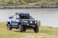 ARB Summit Bumper Kit 3423160KB, off-road vehicle equipped with heavy-duty bumper, tow hooks, and auxiliary lights, parked by a lake on grassy terrain.