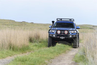 ARB Summit Bumper Kit 3423160KB, black heavy-duty off-road bumper with mounted LED lights and a roof rack on a blue SUV driving off-road in grassy terrain.