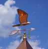 Stunning handcrafted copper peregrine falcon weathervane in mid-flight pose, perched atop a rooftop cupola under a bright blue sky with fluffy clouds. This elegant bird of prey design features detailed wings, a stooping head, and directional arrows marked N, S, E, W for accurate wind indication. Ideal for barns, homes, or gardens—durable, weather-resistant falcon weather vane perfect for bird enthusiasts and rustic decor.

