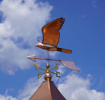 Stunning handcrafted copper peregrine falcon weathervane in mid-flight pose, perched atop a rooftop cupola under a bright blue sky with fluffy clouds. This elegant bird of prey design features detailed wings, a stooping head, and directional arrows marked N, S, E, W for accurate wind indication. Ideal for barns, homes, or gardens—durable, weather-resistant falcon weather vane perfect for bird enthusiasts and rustic decor.

