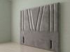 A front-facing view of a headboard upholstered in grey velvet, featuring distinct vertical paneling and a stylized central geometric "W" design created with deep fabric pleats against a plain wall.