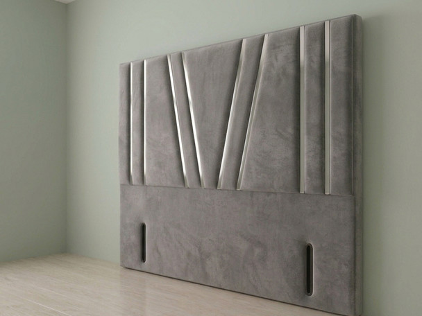 A front-facing view of a headboard upholstered in grey velvet, featuring distinct vertical paneling and a stylized central geometric "W" design created with deep fabric pleats against a plain wall.