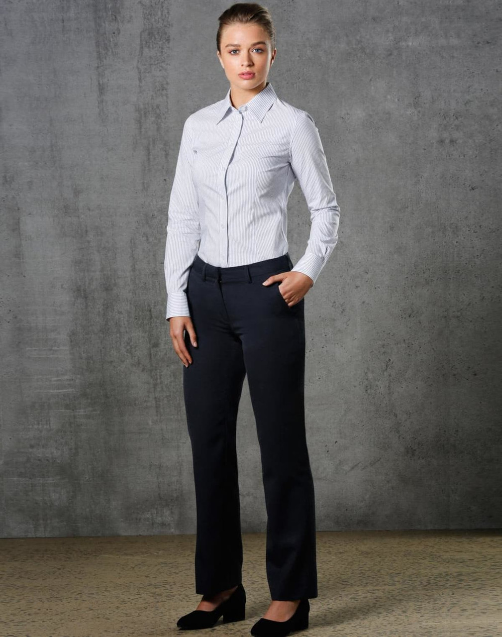 A model wearing ladies' permanent press pants in black, paired with a light blue button-up shirt, standing against a textured backdrop.