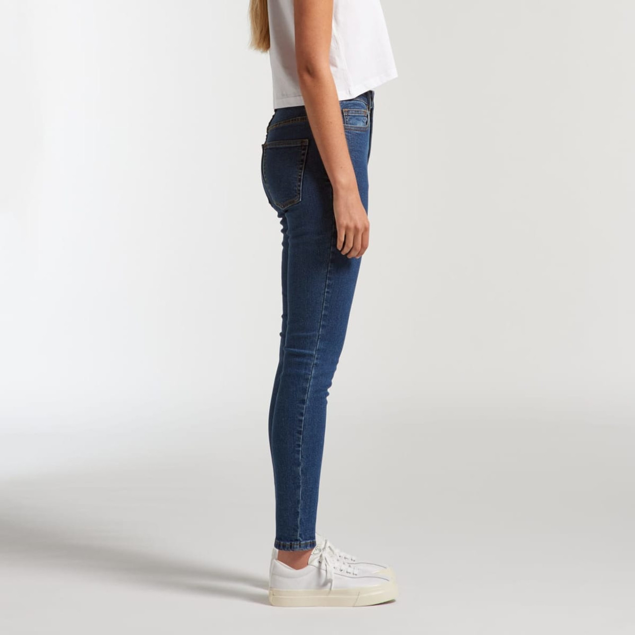 Skinny blue jeans displayed on a model from the side, wearing a white cropped top and white sneakers.