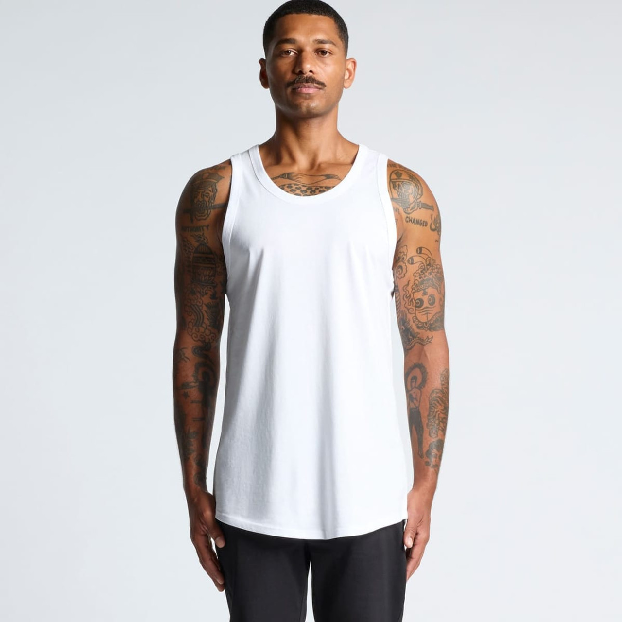 A man wearing a white sleeveless singlet, featuring tattoos on his arms, standing against a light background.