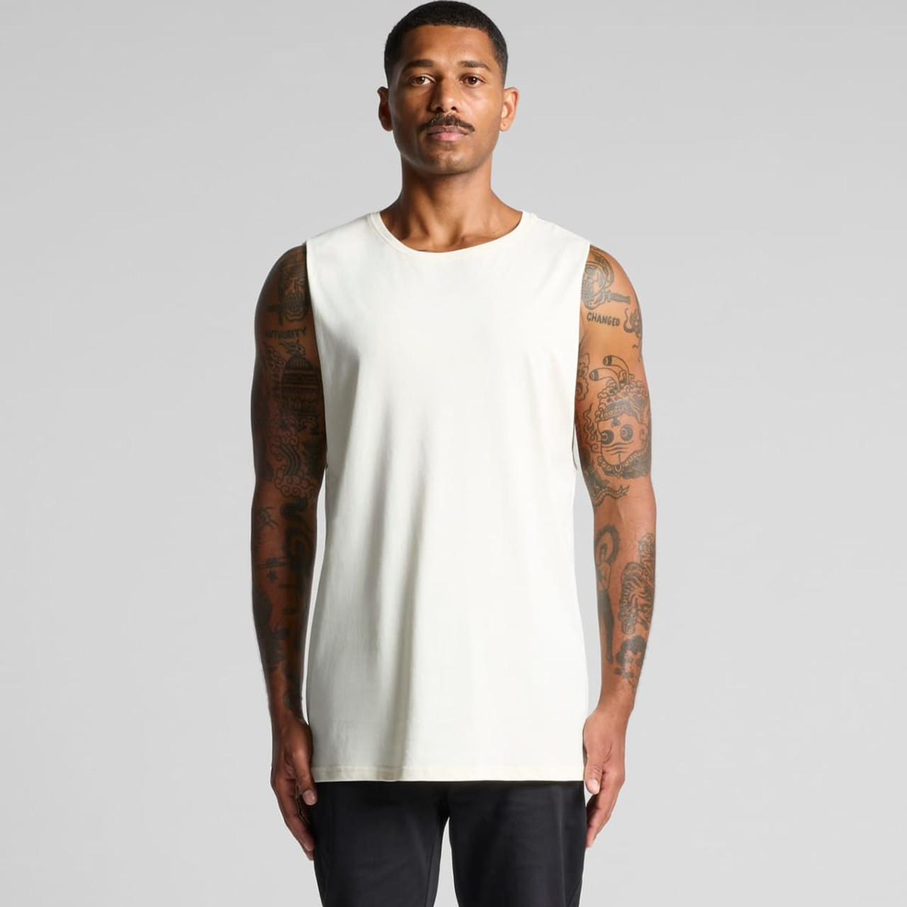 A plain light-coloured singlet tank top worn by a man, featuring a logo on it. The model has tattoos.