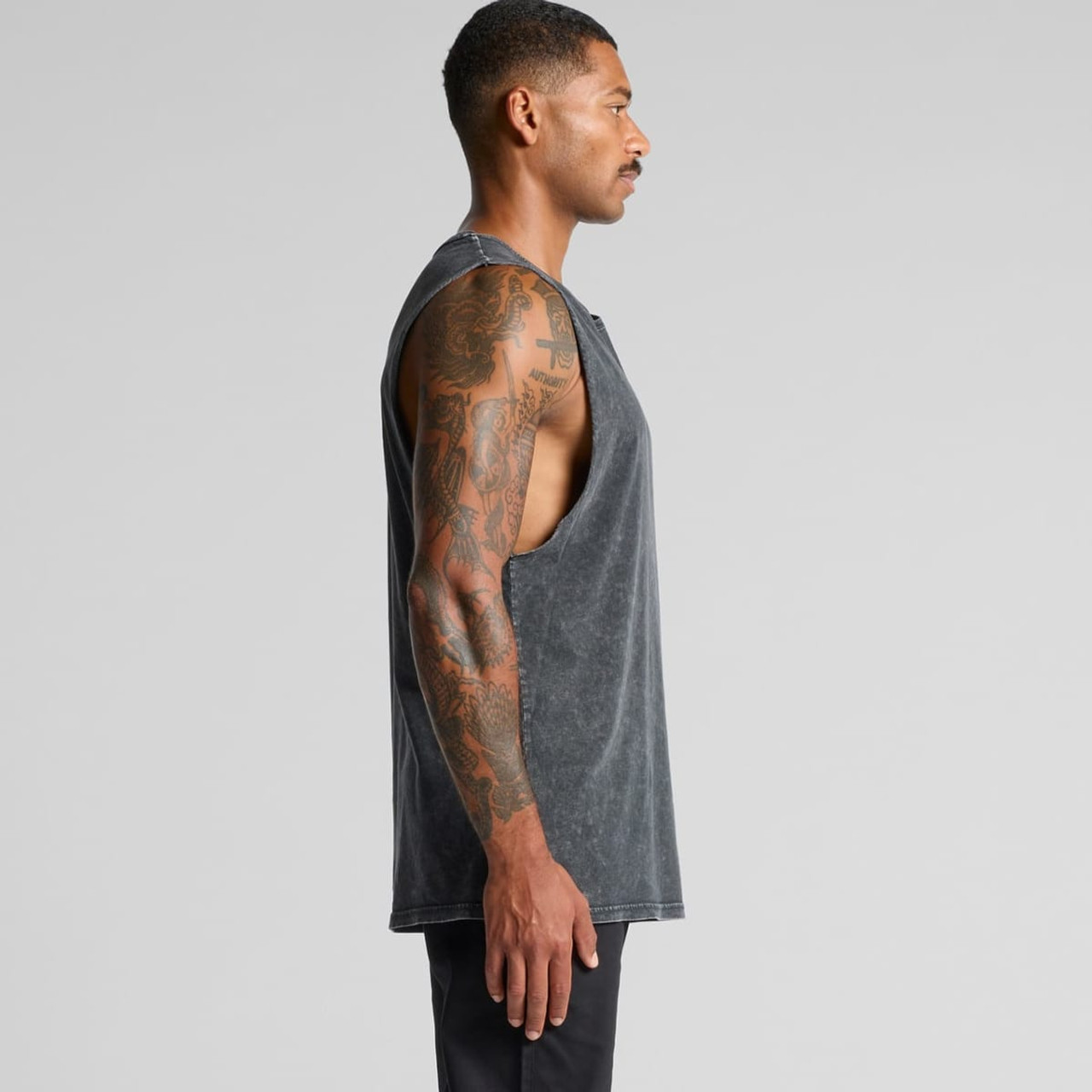 A man with tattoos wearing a grey Stone Wash Barnard Tank top, viewed from the side. The tank has a relaxed fit and sleeveless design.