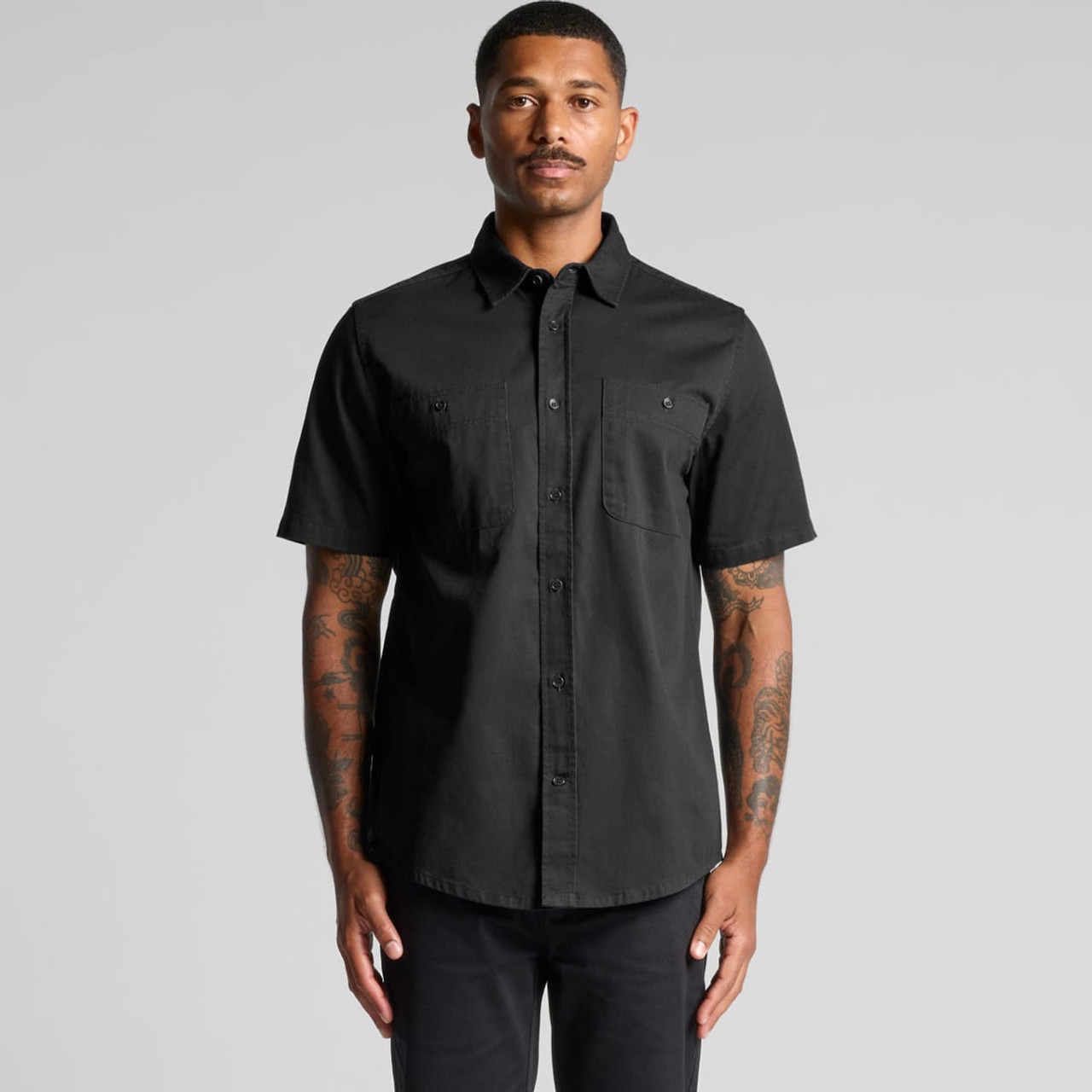 A short-sleeved black work shirt displayed on a model with tattoos, featuring two front pockets and a logo.
