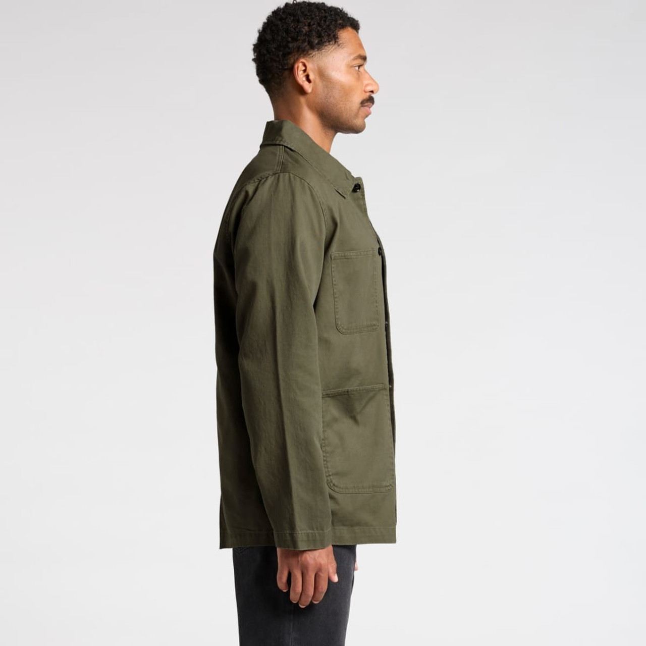 A man wearing an olive green canvas chore jacket, viewed from the side, with a simple design and a logo present.