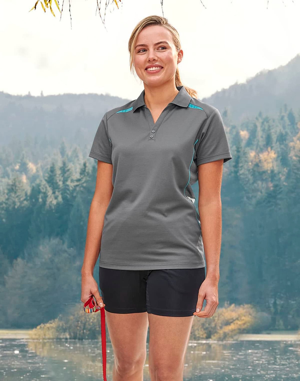 A woman stands outdoors wearing a grey polo shirt with blue contrast accents, alongside black shorts and holding a leash.