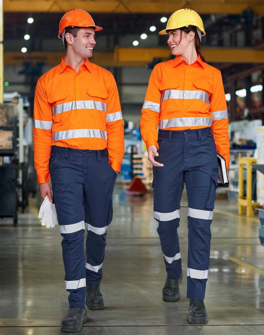 Two workers wear bright orange shirts and navy blue segmented work pants with reflective stripes in an industrial setting.