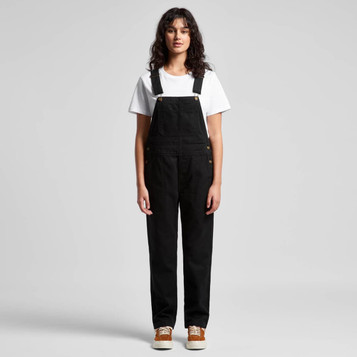 A pair of black canvas overalls worn by a model, paired with a white t-shirt and brown shoes.