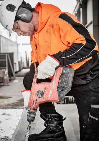 A worker in an orange and black hi-vis pullover hoodie using a power drill while wearing protective gear and gloves.
