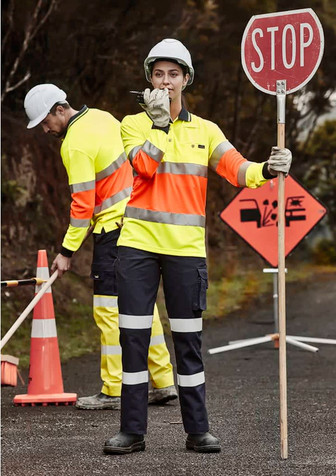 A woman in high-visibility workwear holds a stop sign, while a man in similar gear works nearby. Both wear hard hats.