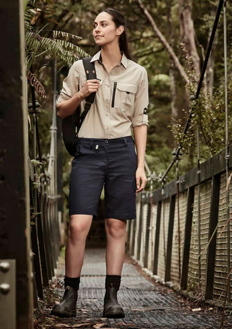 A woman stands on a wooden bridge wearing khaki and navy rugged cooling vented shorts, paired with a button-up shirt and boots.