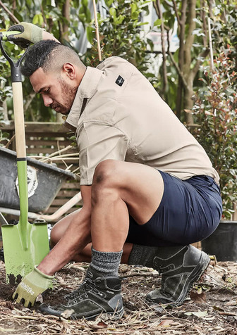 A man in a beige shirt and navy shorts kneels in a garden, working with a green shovel and wearing gloves and boots.