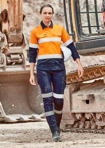 A woman in an orange and navy work outfit with reflective stripes stands near heavy machinery. The outfit includes cargo pants.