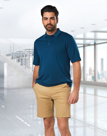 Men's stretch cotton chino shorts in khaki, paired with a navy polo shirt. The model stands in a modern indoor setting.