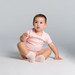 A pink Organic Infant Mini-Me One-Piece displayed on a seated toddler, featuring a simple design and soft fabric.