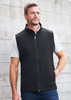 A man models a black vest with a zip front and side pockets against a rustic background. The vest has a logo.