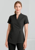 A woman is wearing a black spa tunic with a wrap design and short sleeves. The tunic features a simple button detail.