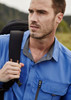 A man wearing a blue short sleeve button-up outdoor shirt, carrying a backpack, with a scenic background.