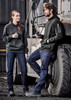 Two individuals wearing black hoodies and blue stretch denim work jeans, standing by a large vehicle.