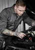 A man in a grey service overall works on a car engine, showcasing detailed tattoos on his arms. The image features a logo on the overall.