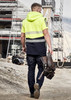 A man in a high-visibility yellow and navy softshell jacket walks at a construction site, holding a tool belt.