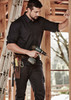 A man in a black long sleeve shirt holds a drill, standing in a wooden structure with a ladder in the background.
