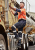 A man in an orange and navy work shirt climbs onto a truck, wearing dark blue stretch jeans with reflective stripes.