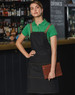 A denim bib apron in dark denim with tan straps, worn by a model holding a clipboard, set against a bar background.