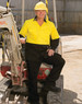 A man wearing a bright yellow and black long-sleeve safety shirt stands beside construction equipment. The shirt features a logo.