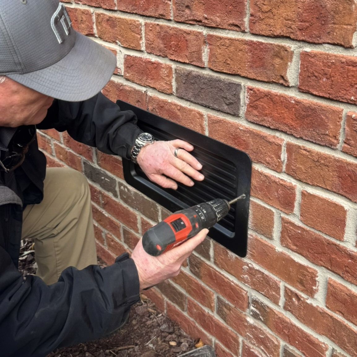 Technician drilling into a mortar joint to prepare for installing an exterior foundation vent cover using included Christmas tree fasteners