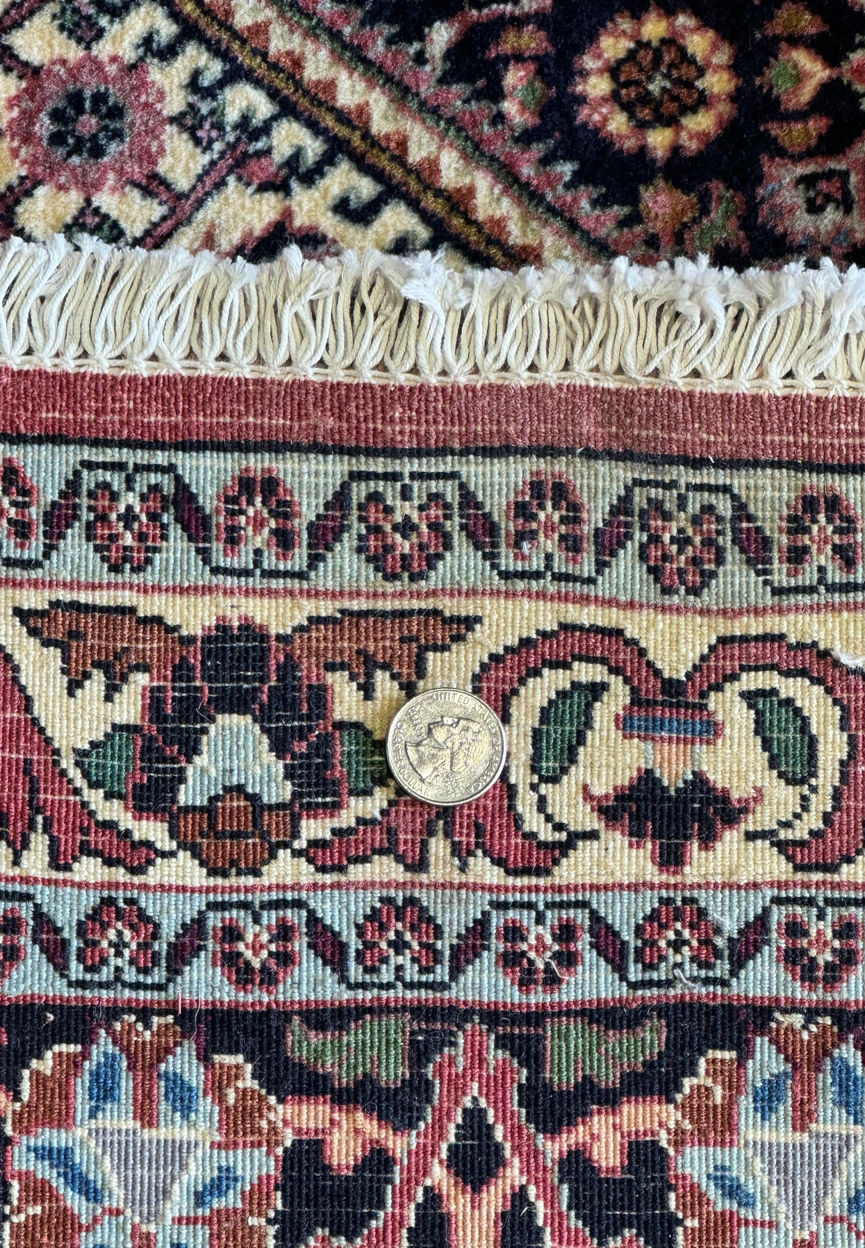 Close-up of a quarter on the Bijar Persian rug's backside, providing a scale reference to the tightly knotted weave. The coin sits atop a complex design featuring detailed flowers and intricate geometric shapes, with the vibrant colors and the knot density highlighting the rug's exceptional quality