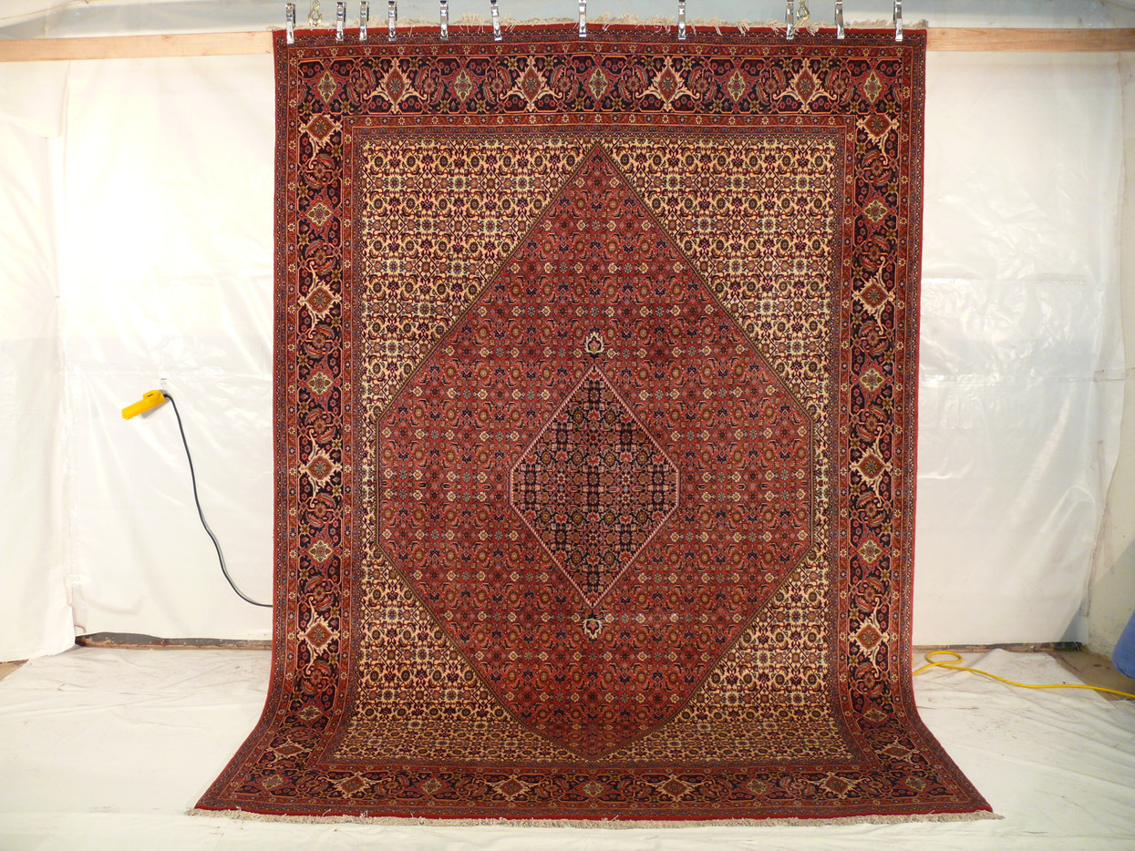 The Persian Bijar Iron Rug hanging vertically, revealing its full size and the dramatic impact of its traditional design against a plain white backdrop, with a yellow extension cord lying on the floor.