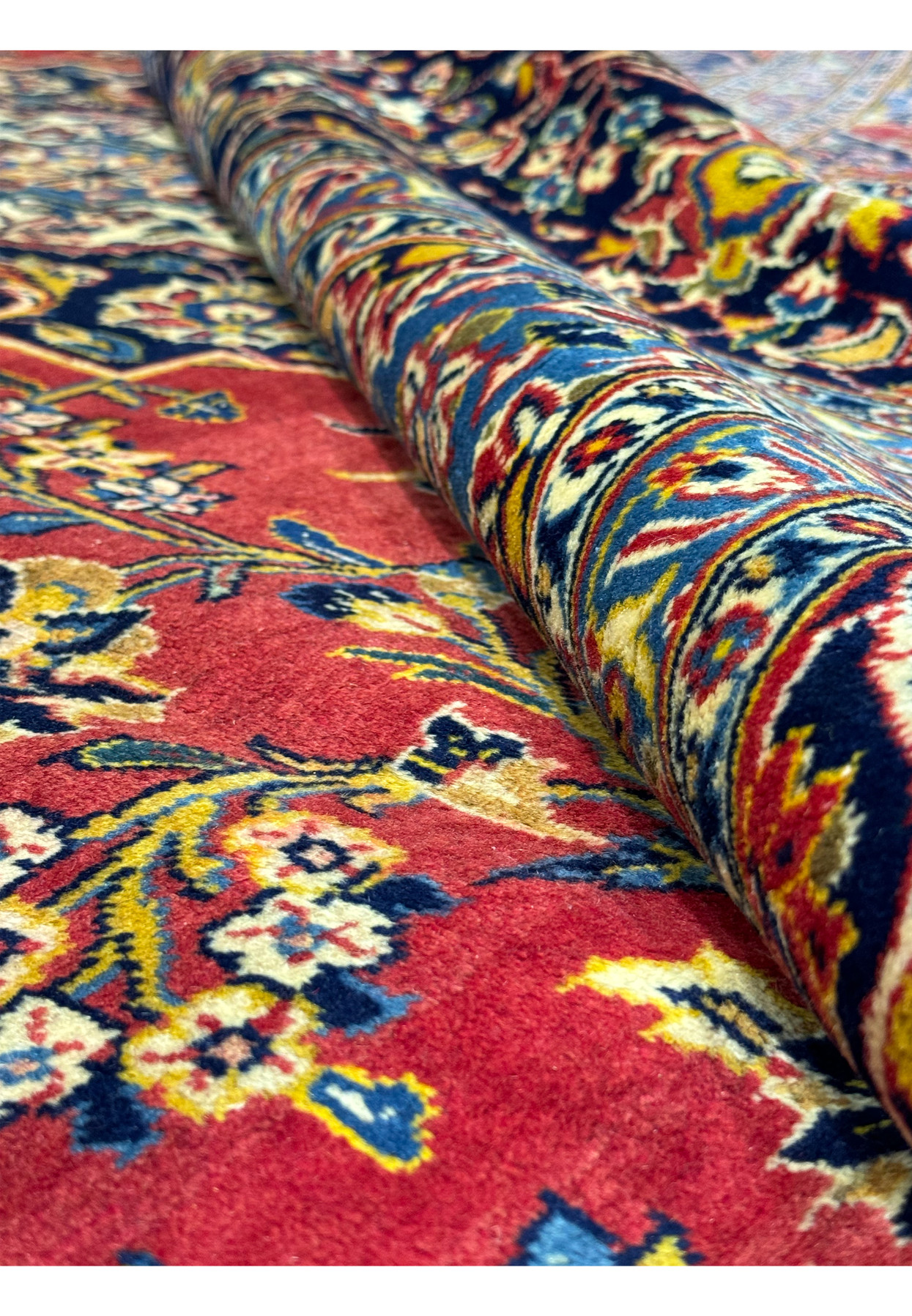Perspective view of a Persian Kashan rug, showing the luxurious texture and detailed design. The rug is partially rolled to reveal the intricate border and floral patterns against a red field. The rich colors and high-quality materials are evident, making this rug a stunning addition to any room. This image emphasizes the durability and elegance of Kashan rugs.